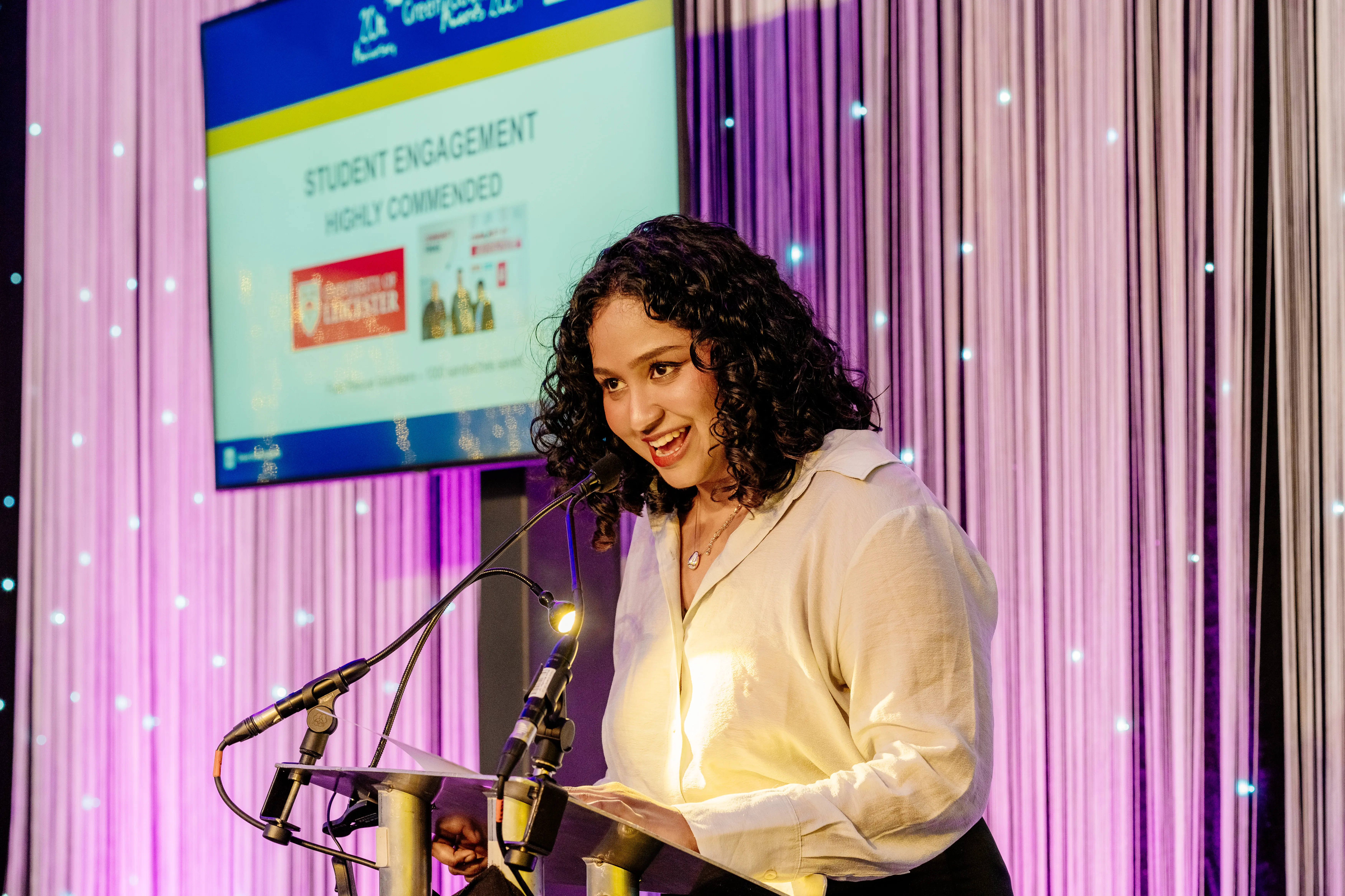 A woman with long curly hair and white blouse stands at a lectern with a large screen in the background and glittery backdrop. 